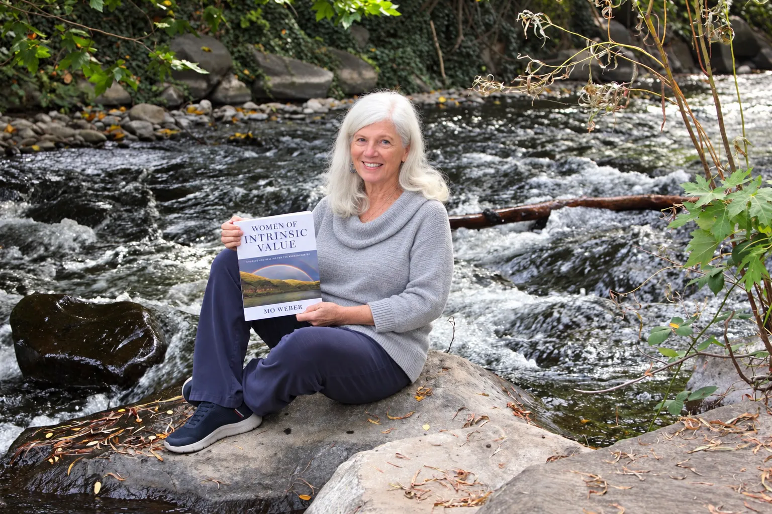 Mo Weber sitting on a rock beside a rushing river, smiling and holding a copy of Women of Intrinsic Value